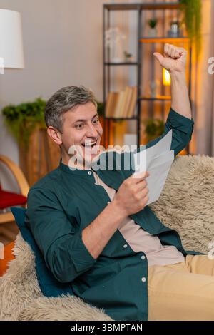 Junger glücklicher Mann zu Hause öffnet den Brief und feiert glücklich große Nachrichten, indem er vor Freude die Faust erhebt. Der Kaukasier auf dem Sofa ist begeistert von Stellenangeboten, Kreditgenehmigungen oder erfolgreichen Schulprüfungen. Stockfoto