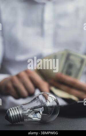 Elektrische Glühlampe. Der Mann übergibt Zählkosten für Elektrizitätsscheine und Bargeldbuchungen in Dollar an den Rechner. Nahaufnahme der Hände, die nicht erkennbar sind Stockfoto