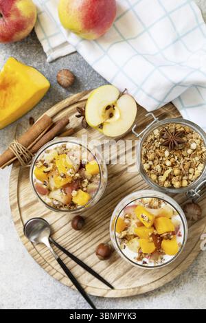 Gesundes Frühstücksgranola im Herbst oder Winter. Hausgemachtes Dessert mit Joghurt, Müsli, Karamelläpfeln und Kürbis, Zimt, Nüssen auf einem Steintisch. Ansicht f Stockfoto