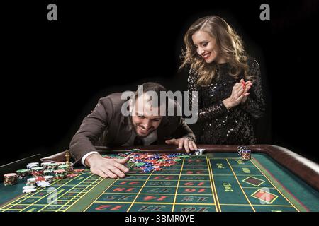 Ein Mann in einem braunen Anzug mit einer schönen Frau in einem schwarzen Kleid spielt an einem Roulette-Tisch in einem Casino. Emotionen von Glücksspielern Stockfoto