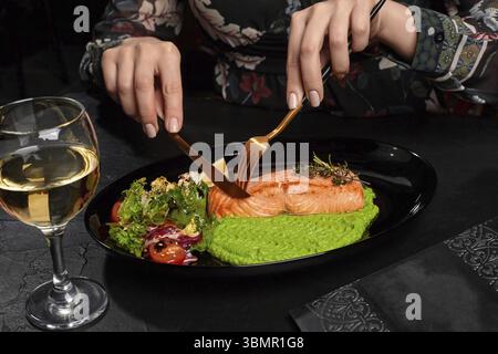 Weibliche Hände schneiden appetitlich gegrilltes Lachssteak auf dem Teller serviert mit grünem Erbsenpüree, frischen Kräutern und Gemüsesalat, Zitronenscheibe und Glas Stockfoto