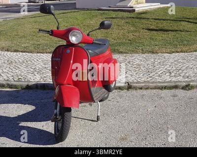 Glänzend roter Vespa-Roller parkt auf einer asphaltierten Straße neben einem grasbewachsenen Gebiet Stockfoto