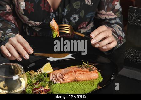 Hände einer jungen Frau, die Messer und Gabel mit einem Stück appetitlich gebackenem Forellensteak mit pürierten grünen Erbsen, frischem Salat, Zitronenscheibe und aro hält Stockfoto