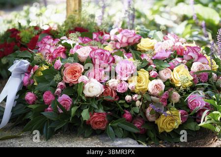 Ein Trauerkranz aus orangen, rosa, gelben und weißen Rosen und einem weißen Band liegt auf einem frischen Grab auf einem Friedhof Stockfoto