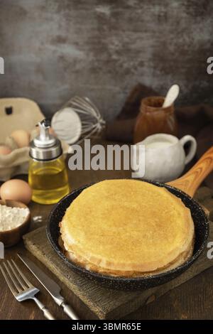 Wir feiern den Pfannkuchen-Tag, kochen ein gesundes Frühstück. Köstliche hausgemachte Crepes oder Pfannkuchen in einer Pfanne und Zutaten auf einem rustikalen Tisch. Kopieren Sie Spac Stockfoto