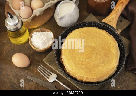 Pancake Day feiern, gesundes Frühstück kochen. Köstliche hausgemachte Crepes oder Pfannkuchen in einer Pfanne und Zutaten auf einem rustikalen Tisch Stockfoto