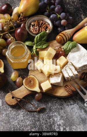 Käseplatte, Stillleben von saisonalen Weinsnacks. Brie-Käse, frische Feigen, Trauben, Käsewürfel, Birnen, Pekannüsse und Honig auf dem Schneidebrett im Dunkeln Stockfoto