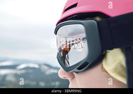 Nahaufnahme des Porträts einer Snowboarderin im Skigebiet, die Helm und Schutzbrille trägt, die Berge reflektiert, und eines anderen Snowboarders oder Skifahrers und Skifahrers Stockfoto
