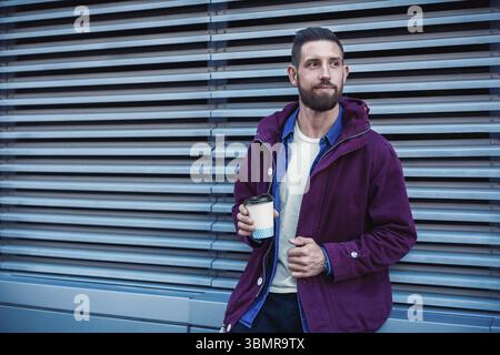Hübscher junger Mann in lila Winterjacke Kaffee zu trinken. Der Kerl trinkt Kaffee auf der Straße. Mann hält Pappbecher mit Americano oder Latte. Stre Stockfoto