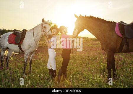 Zwei Frauen und zwei Pferde draußen im Sommer glücklicher Sonnenuntergang zusammen Natur. Tierpflege, Liebes- und Freundschaftskonzept Stockfoto