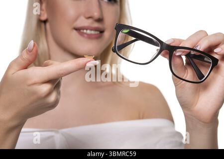 Junge blonde Frau mit Kontaktlinsen auf den Finger vor ihrem Gesicht und halten in der anderen Hand eine schwarze Brille auf weißem Hintergrund. Close-up. Ey Stockfoto