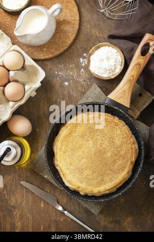 Wir feiern den Pfannkuchen-Tag, kochen ein gesundes Frühstück. Köstliche hausgemachte Crepes oder Pfannkuchen in einer Pfanne und Zutaten auf einem rustikalen Tisch. Ansicht von Stockfoto