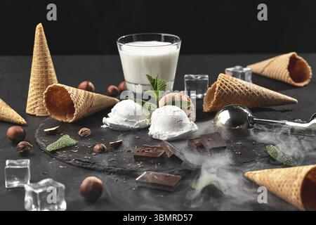 Es werden Schokoladen- und cremige Eiskugeln serviert, die mit Minze dekoriert sind, ein Glas Milch, Eiswürfel, Waffelkegel mit Schokolade und Haselnüssen in der Nähe Stockfoto