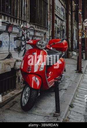 Klassischer roter Vespa-Roller, der auf einem Bürgersteig vor einer mit Graffiti bedeckten Wand geparkt ist und urbanen Stil und Freiheit verkörpert Stockfoto