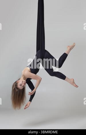 Professionelle Turnerleistung auf schwarzen Leinwänden. Anmutiges Mädchen in einem dunklen Sportanzug macht akrobatische Elemente und lächelt beim Aufhängen von upsi Stockfoto