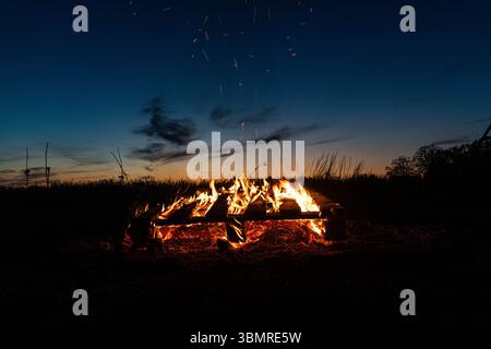 Landkreis Osnabrück, Deutschland 19. April 2025: Im Bild: Ein fesselndes Lagerfeuer brennt mit leuchtenden Flammen und einem beeindruckenden Funkenflug in der Dämmerung oder bei Nacht. Die Holzscheite und Paletten bilden die Basis dieses Feuers, dessen warmes, Orangen Licht die umgebende Landschaft erhellt. Der klare Abendhimmel mit sanften Farbübergängen unterstreicht die ruhige und doch dynamische Atmosphäre. Dieses Bild ist ideal für redaktionelle Inhalte über Outdoor-Aktivitäten, Camping-Erlebnisse, Freizeitgestaltung, romantische Momente in der Natur oder die Darstellung traditioneller Fe Stockfoto