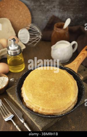 Pancake Day feiern, gesundes Frühstück kochen. Köstliche hausgemachte Crepes oder Pfannkuchen in einer Pfanne und Zutaten auf einem rustikalen Tisch Stockfoto