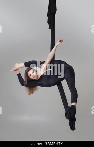 Turnerleistung auf schwarzen Leinwänden. Elegante Dame im dunklen Sportanzug schaut in die Kamera und macht akrobatische Elemente, die an einem Seil hängen Stockfoto