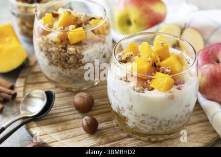 Gesundes Herbst- oder Winterfrühstück. Hausgemachtes Dessert mit Joghurt, Müsli, Karamell-Äpfeln und Kürbis, Zimt, Nüssen auf einem Steintisch Stockfoto