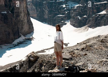 Junge Frau, die auf felsigem Hang über dem Gletscher steht, mit hohen Klippen im Hintergrund Stockfoto