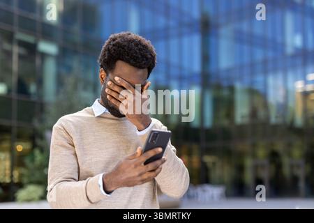 Ein besorgter Mann bedeckt sein Gesicht, scheinbar beunruhigt, während er sein Handy ansieht, vor einem modernen Gebäude Hintergrund. Stockfoto