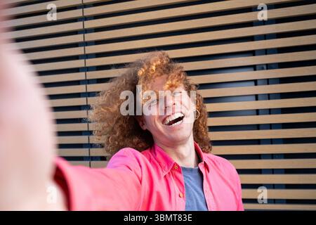 Selfie eines fröhlichen jungen Mannes mit lockigen Haaren, der fröhlich vor einer Holzlattenwand lacht. Mit rosa Hemd, lebhaften Emotionen, lässigem Style, aut Stockfoto