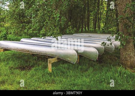 Mehrere Metallkanus stehen kopfüber auf Holzregalen im schattigen Bereich in der Nähe des Waldes, bereit für Sommeraktivitäten. Schweden. Stockfoto