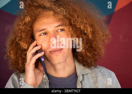 Nahaufnahme eines jungen Schülers mit lockigen Haaren, der auf dem Smartphone spricht, nachdenklicher Ausdruck vor einem farbenfrohen Wandgemälde. Urbane Umgebung, Universitätsleben Stockfoto