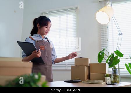 Junge asiatische Frau in Schürze, die von zu Hause aus Aufträge für den Versand vorbereitet. Sie verpackt Kartons für die Lieferung in ihrem Online-Shop, der einen E-Commerce betreibt Stockfoto