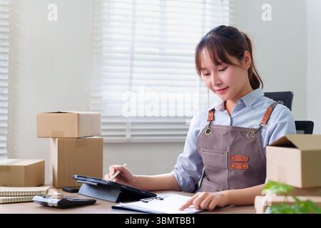 Eine junge asiatische Frau, die eine Schürze trägt, nutzt ein Tablet, um ihr Online-Geschäft von zu Hause aus zu verwalten, während sie von Paketfächern umgeben ist. Sie sieht glücklich und org aus Stockfoto