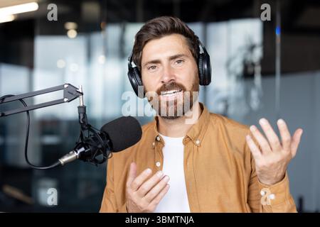 Ein Mann, der Kopfhörer trägt, spricht in einem Podcast-Studio in ein Mikrofon und bewegt mit seinen Händen. Stockfoto