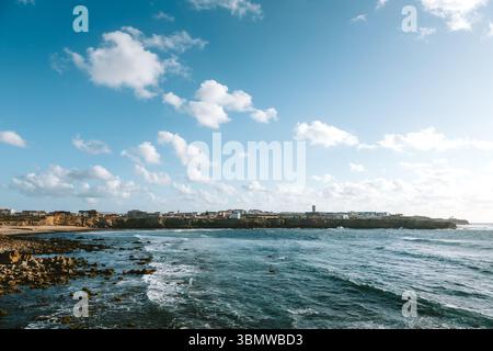 Malerische Küstenszene in Peniche, Portugal. Wellen krachen gegen die felsige Küste, mit der Stadtlandschaft im Hintergrund unter einem bewölkten Himmel. Stockfoto