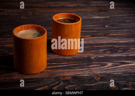 Zwei Keramikbecher, eine mit schaumigem Espresso und die andere mit schwarzem Kaffee, sitzen nebeneinander auf einem rustikalen Holztisch in gemütlicher und natürlicher Umgebung Stockfoto