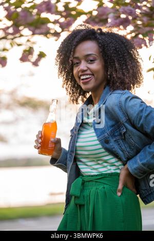 Eine junge afroamerikanische Frau mit natürlich lockigem Haar trinkt an einem warmen Tag erfrischende Orangensoda. Stockfoto