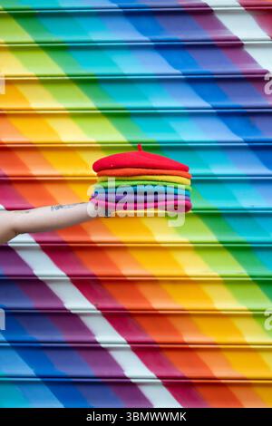 Eine junge Frau hält einen Stapel farbiger Wollmützen, während sie vor einem Regenbogengemälde steht. Stockfoto