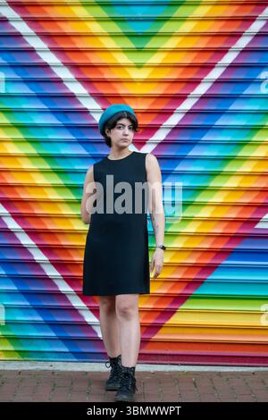 Eine junge Frau posiert vor einem Regenbogengemälde mit einer bunten Baskenmütze. Stockfoto