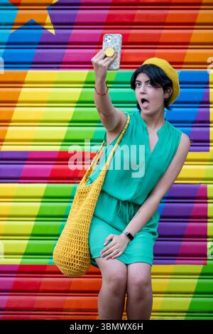 Eine junge Frau in grünem Kleid und gelber Baskenmütze macht Selfies vor einem Regenbogengemälde. Stockfoto