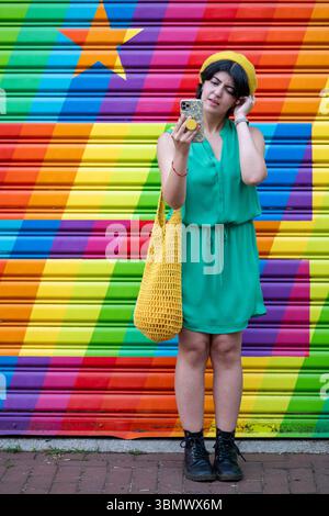 Eine junge Frau in grünem Kleid und gelber Baskenmütze macht Selfies vor einem Regenbogengemälde. Stockfoto