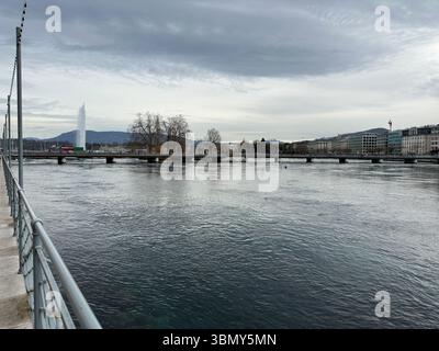 Genf, Schweiz - 12. Januar 2023. Der ikonische Jet d'Eau-Brunnen erhebt sich über dem ruhigen Wasser des Genfer Sees mit einer Stadtbrücke. Stockfoto