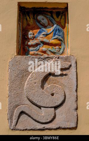 Reliefschnitzerei auf Steintafel, wahrscheinlich mittelalterlich, von gewickelter Schlange oder Schlange, mit offenem Mund und spitzen Ohren. Eingebettet in die Hauswand in der ehemaligen etruskischen und römischen Stadt Toskana in der Provinz Viterbo, Latium, Italien, unter der glasierten und bemalten Terrakotta-Darstellung von Madonna und Kind. Stockfoto