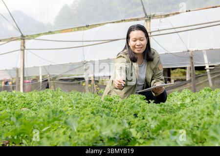 Asiatische Farmerin, die mit Freude Daten zur Gemüseanbaufläche aufzeichnet, repräsentiert nachhaltiges Agrarunternehmen. Stockfoto
