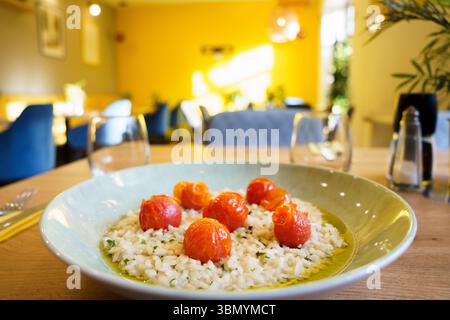 Ein Teller mit cremigem Risotto, garniert mit gerösteten Kirschtomaten, serviert auf einem Holztisch in einem warm beleuchteten Restaurant. Die leuchtend gelbe Wand und das weiche Fu Stockfoto