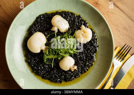 Ein Gourmet-Teller mit Risotto aus schwarzer Tintenfisch-Tinte, garniert mit gebratenem Tintenfisch und einer frischen Rucola-Garnitur. Der leuchtende Grün- und Tiefschwarz-Kontrast b Stockfoto