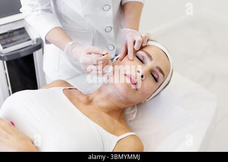 Reife Frau bekommt Verjüngungsspritze in Schönheitsklinik durch Kosmetikerin in Schutzhandschuhen und weißem Fell, Anti-Aging-Gesichtsbehandlung zum Smoo Stockfoto