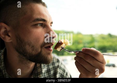 Ein bärtiger Mann, der gerade einen Bissen Heidelbeerkuchen essen will, hält die Gabel neben seinen Mund und blickt nach vorne in ein helles, natürliches Café. Schließen Sie Seitenportren Stockfoto
