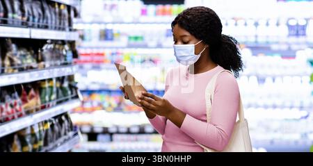 Millennial schwarze Dame mit Tragetasche trägt Gesichtsmaske, Einkaufen für Lebensmittel im Einkaufszentrum während covid Ausbruch Stockfoto