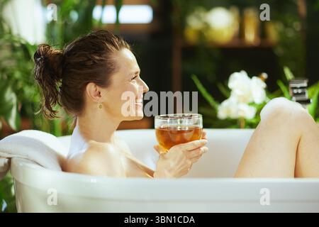 Eine Frau mit nassem Haar und entspanntem Lächeln ist im Seitenprofil festgehalten und genießt ein wohltuendes Schaumbad in einer weißen Wanne mit Tee. Verschwommene grüne Pflanzen Stockfoto
