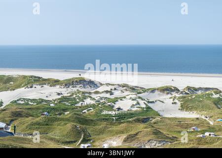 Weitläufige Sanddünen erstrecken sich entlang des Strandes, umgeben von tiefblauem Meer. Besucher spazieren entlang der Küste unter der hellen Mittagssonne und genießen die Stockfoto