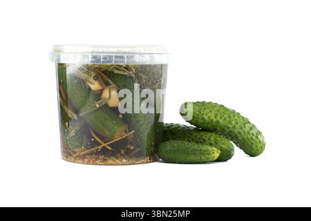 Eingelegte Gurken in einem transparenten Plastikglas mit frischen Kräutern und Gewürzen, zusammen mit rohen Gurken, dargestellt auf einem stark weißen Hintergrund, Betonung Stockfoto
