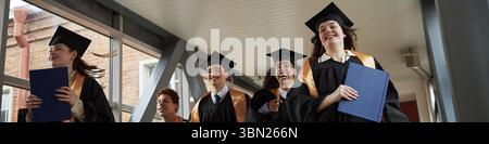 Kopfzeile der Gruppe multiethnischer junger Menschen, die Abschlussmützen und -Kleider tragen, die zusammen laufen, Diplome halten, lächeln und akademische Leistungen in der Universität feiern Stockfoto